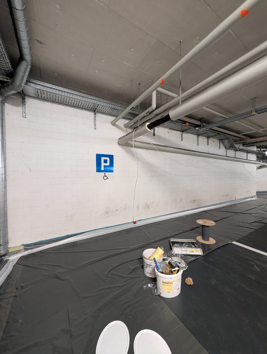 Tiefgarage mit weiss gestrichener Wand, Rohrleitungen an der Decke und abgedecktem Boden